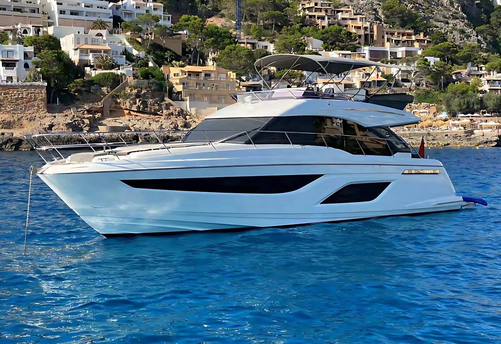 Bavaria R55 flybridge yacht at anchor in a blue bay below hillside villas and pine trees