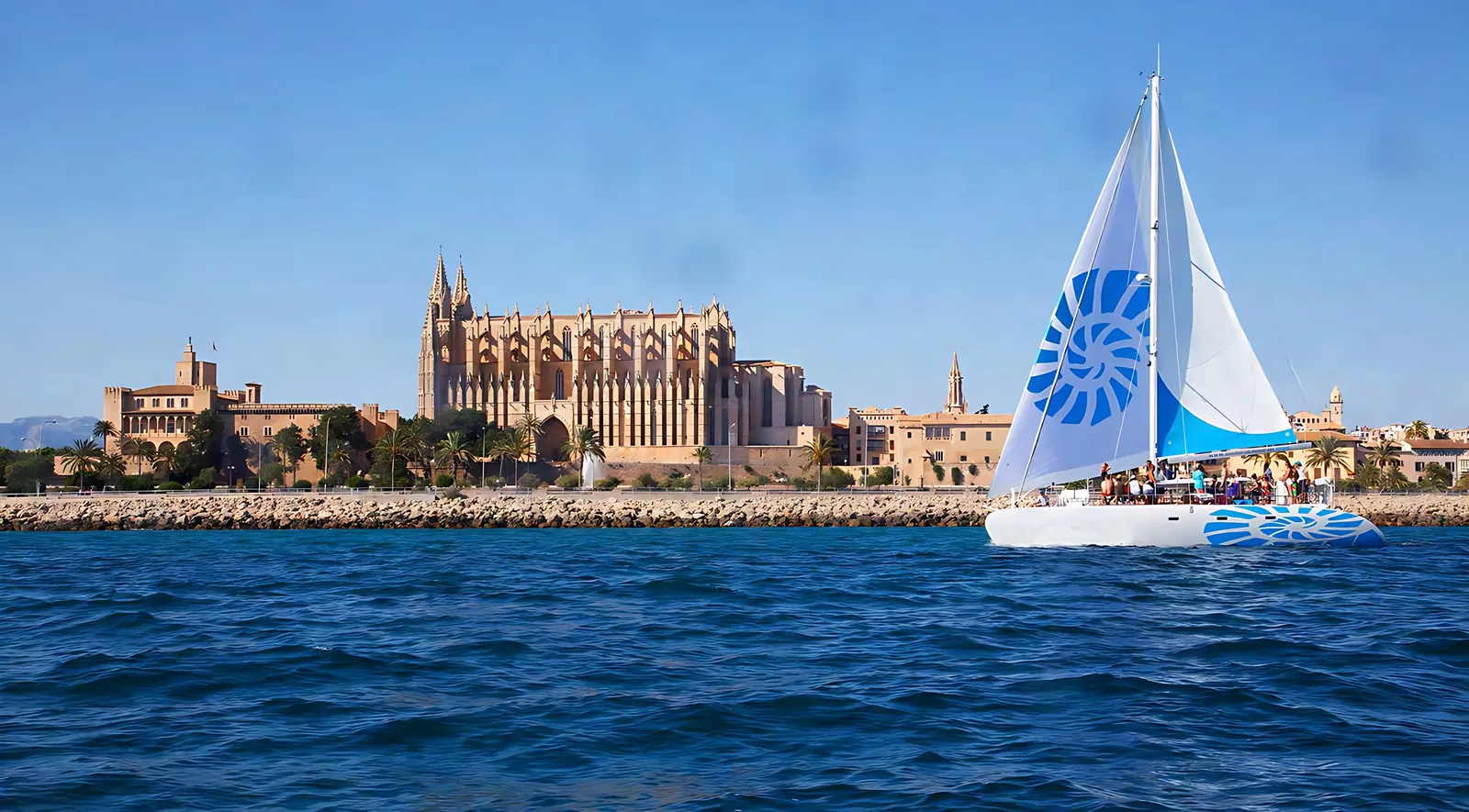 Catamaran under sail with guests on deck, passing Palma Cathedral and the Almudaina palace on the Bay of Palma