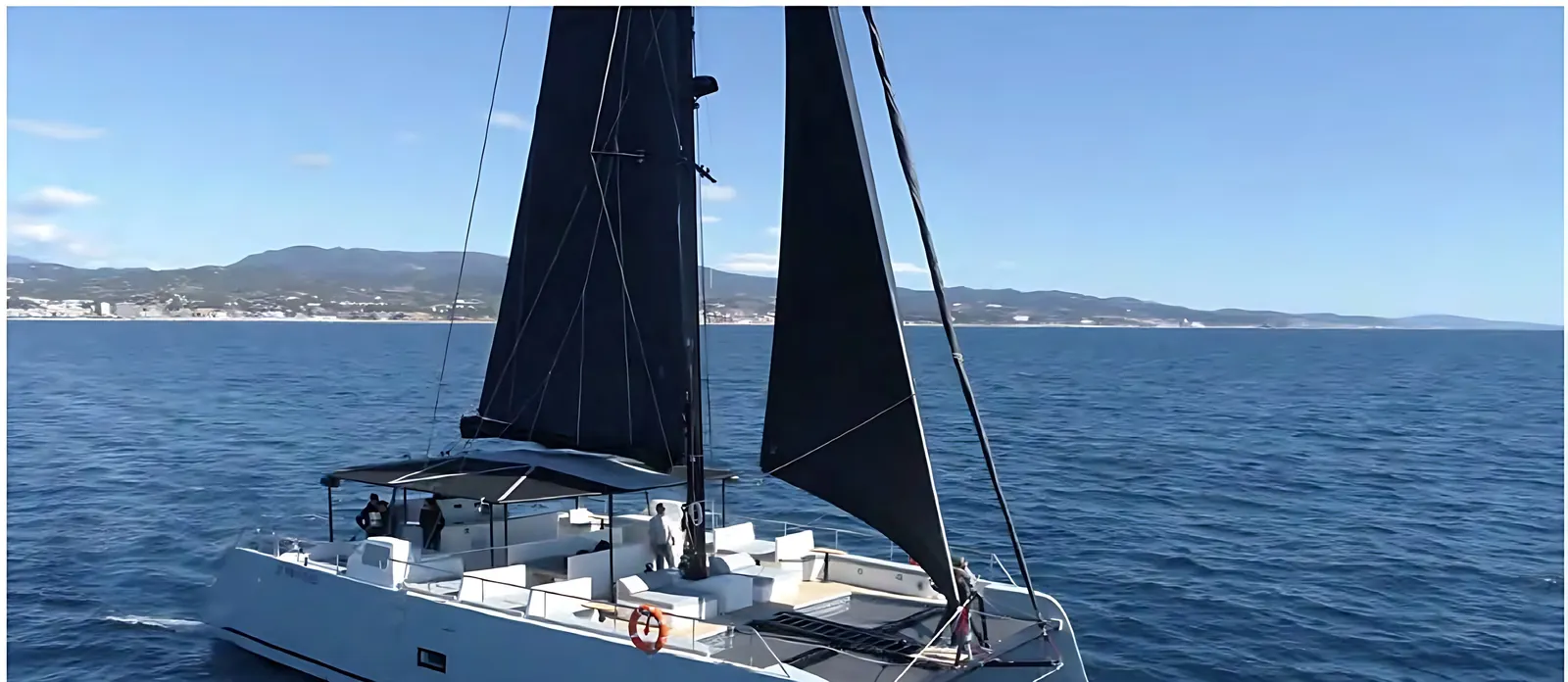 Catamaran II under dark sails off a hilly coastline, guests on her wide deck beneath a shade canopy