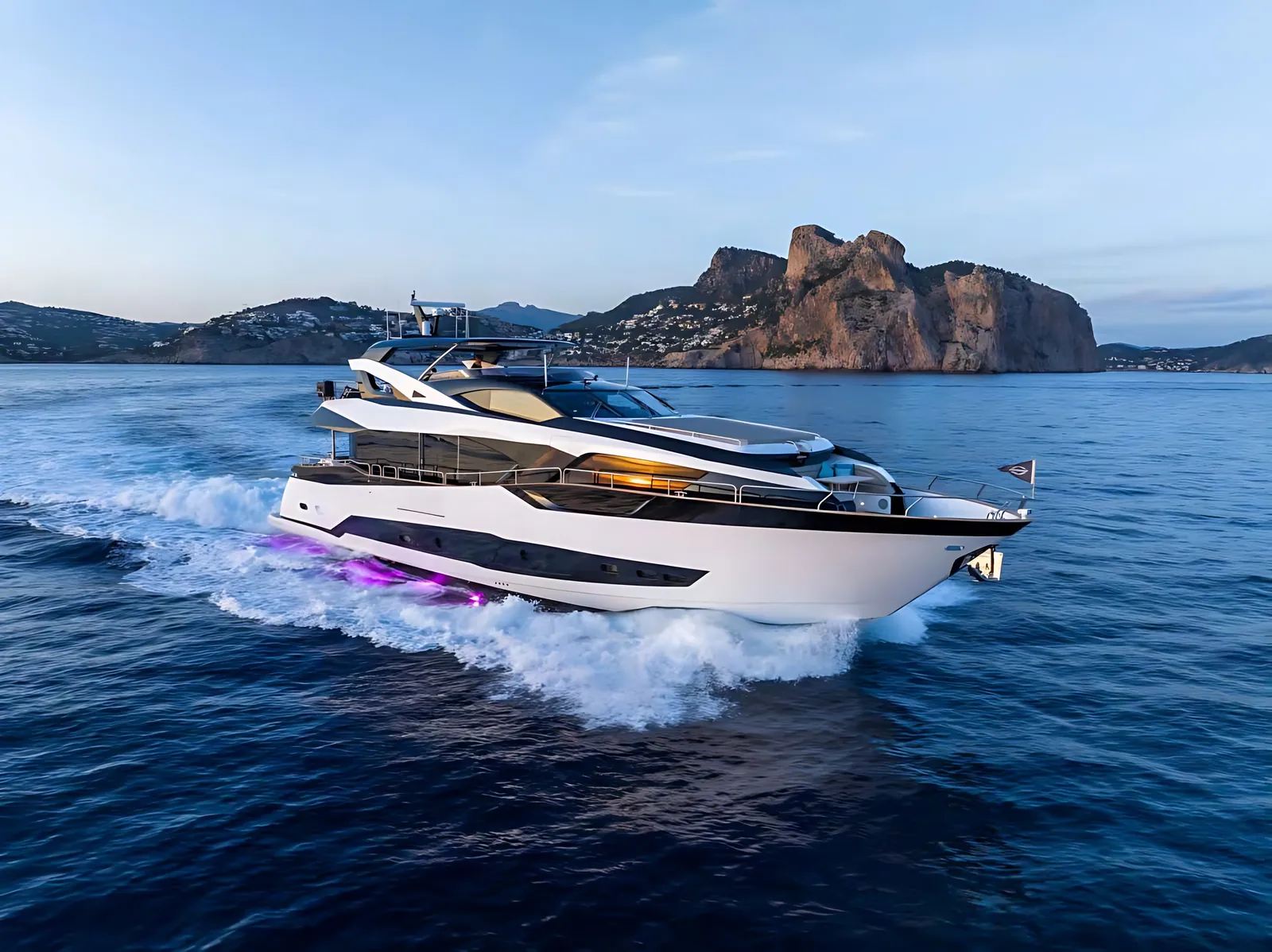 Sunseeker 95 flybridge yacht under way at dusk, white wake behind and a rocky headland in the distance